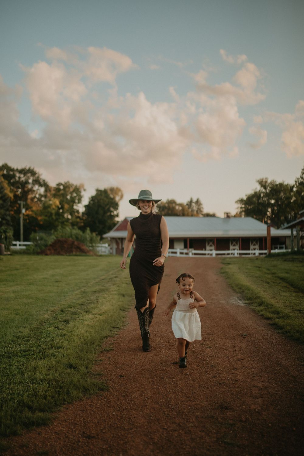Two people walk together on a dirt path surrounded by green grass and trees under a cloudy sunset sky.