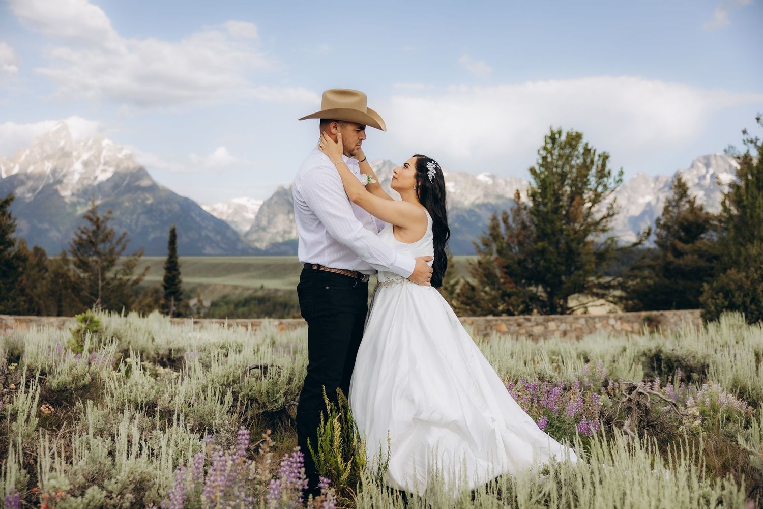 A couple in wedding attire poses against a stone wall with majestic snow-capped mountains in the background.