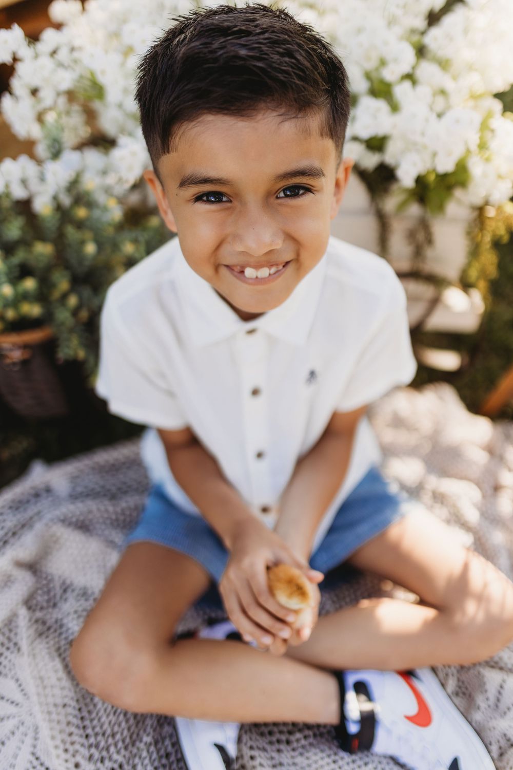 Smiling young boy in white shirt and denim shorts sitting cross-legged outdoors near white flowers.