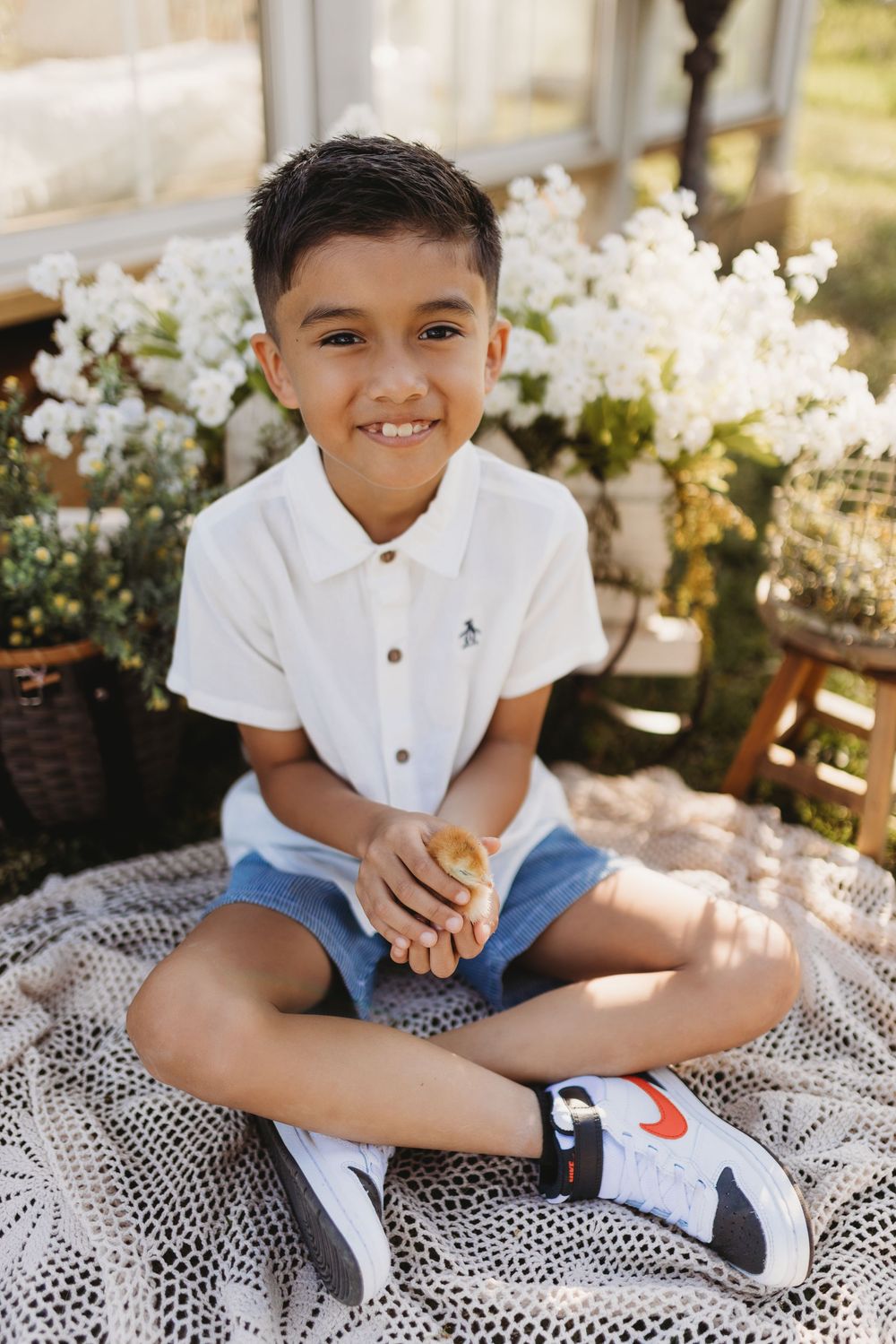 Smiling boy in white polo shirt and denim shorts sitting cross-legged on blanket with white flowers in background.