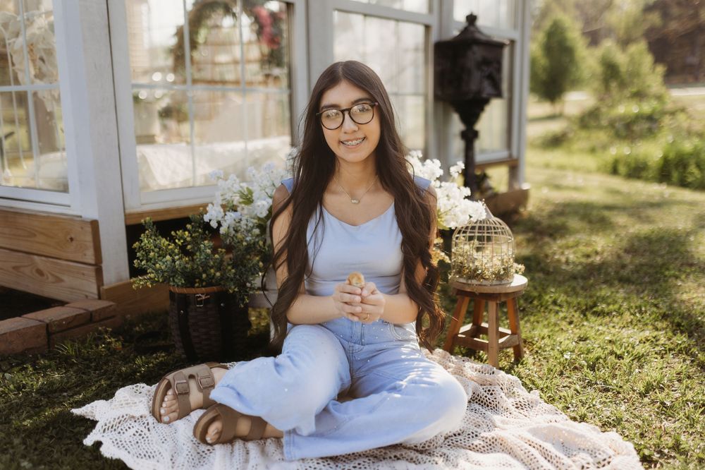 Young woman in blue outfit sitting cross-legged on blanket in garden near greenhouse with flowers.