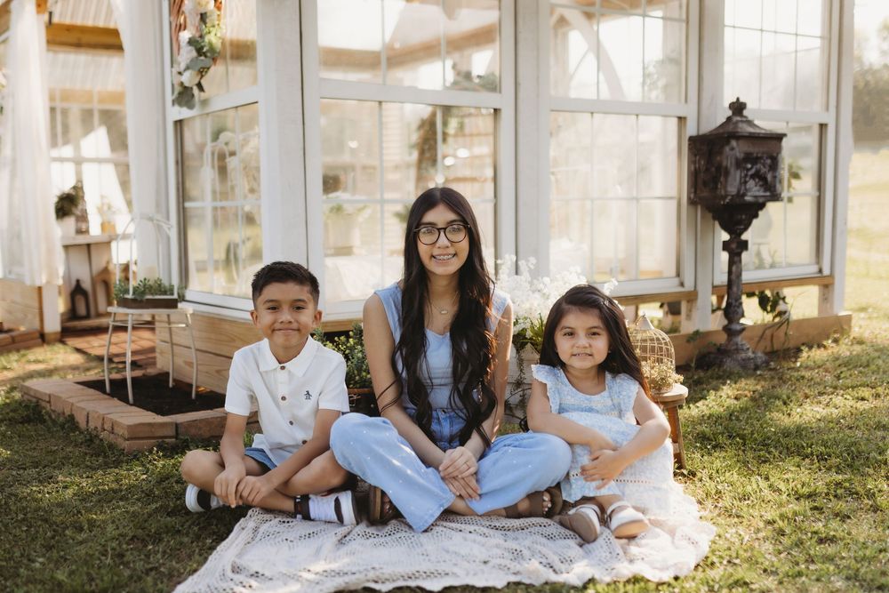A woman with glasses sits cross-legged on a blanket outdoors with two young children in front of a greenhouse.