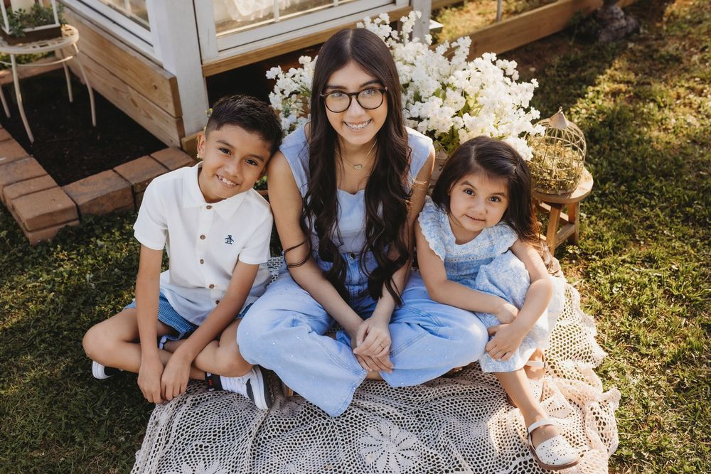 Woman with glasses and two young children sitting on a blanket outdoors near white flowers.
