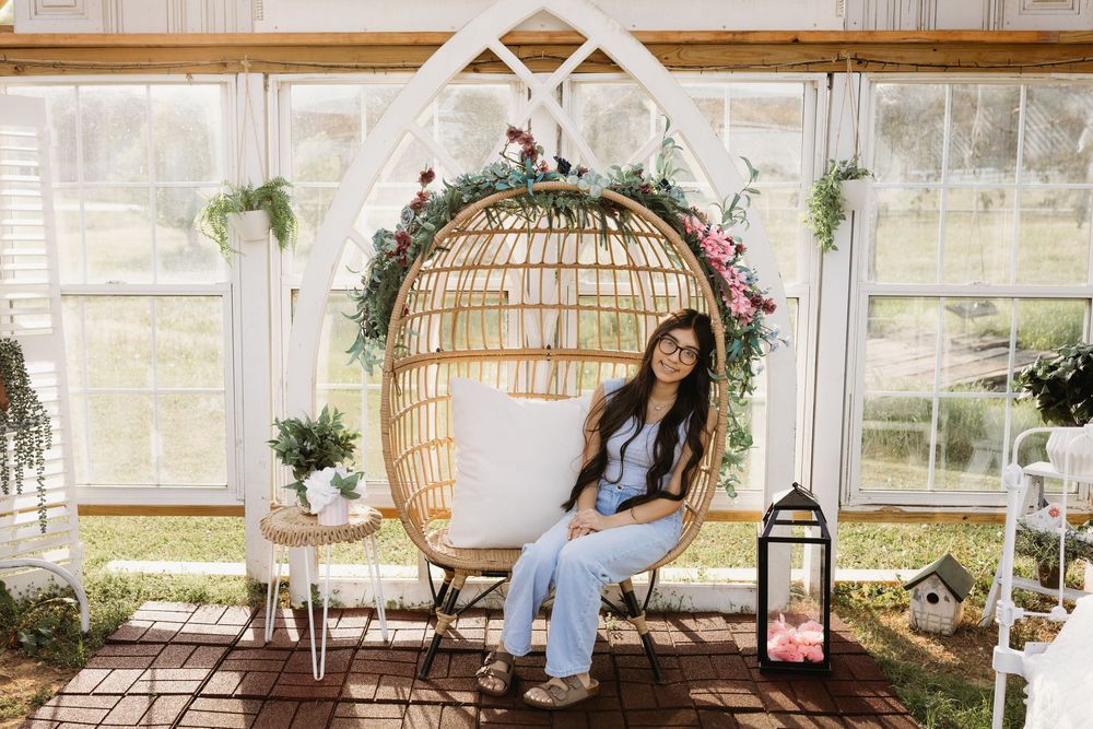 Woman sitting in a rattan egg chair decorated with flowers inside a bright greenhouse setting.