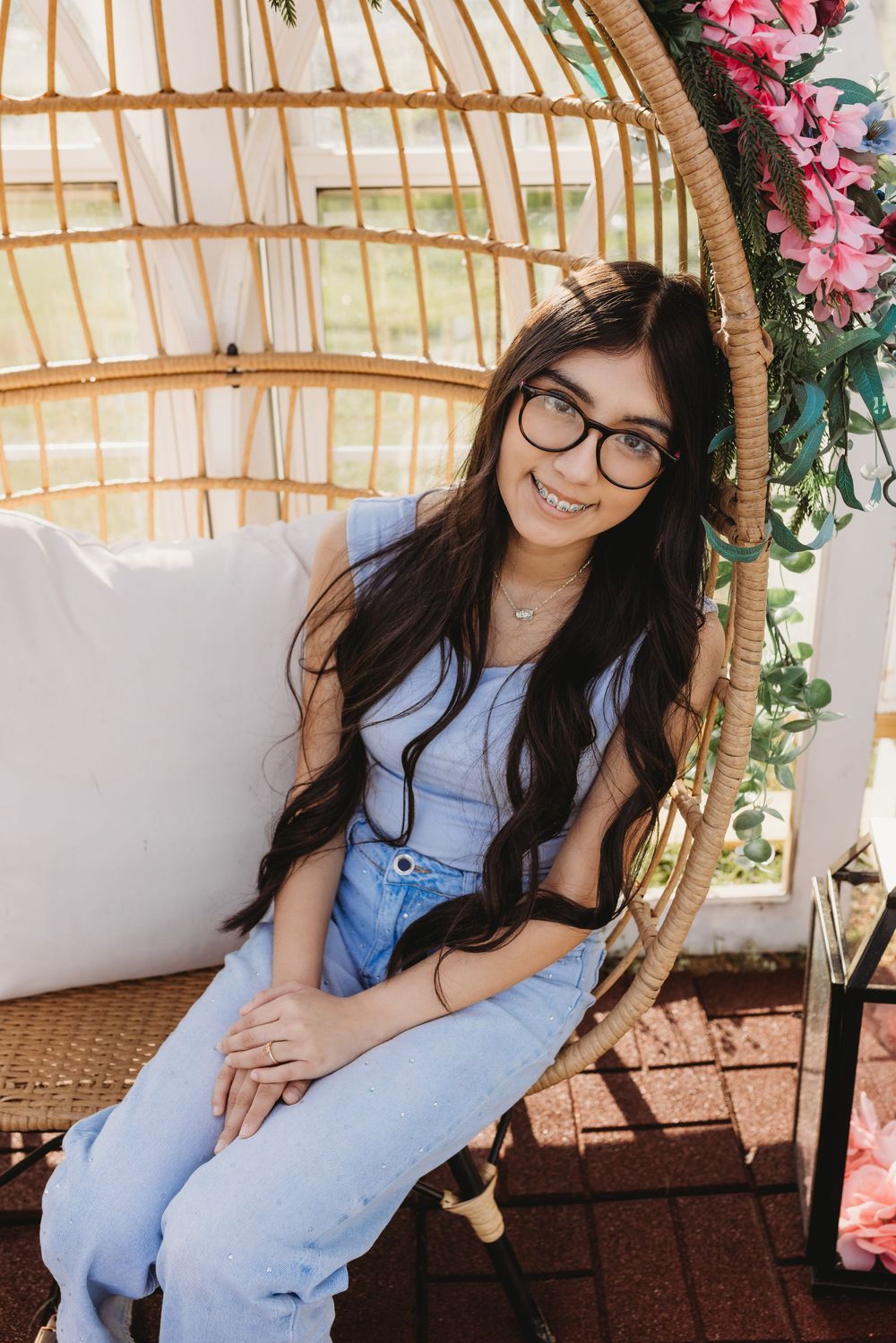 Young woman with long dark hair and glasses smiling in rattan egg chair with pink flowers.