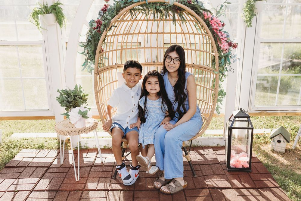 A woman and two children smile together in a wicker egg chair decorated with floral greenery in a bright, airy space.