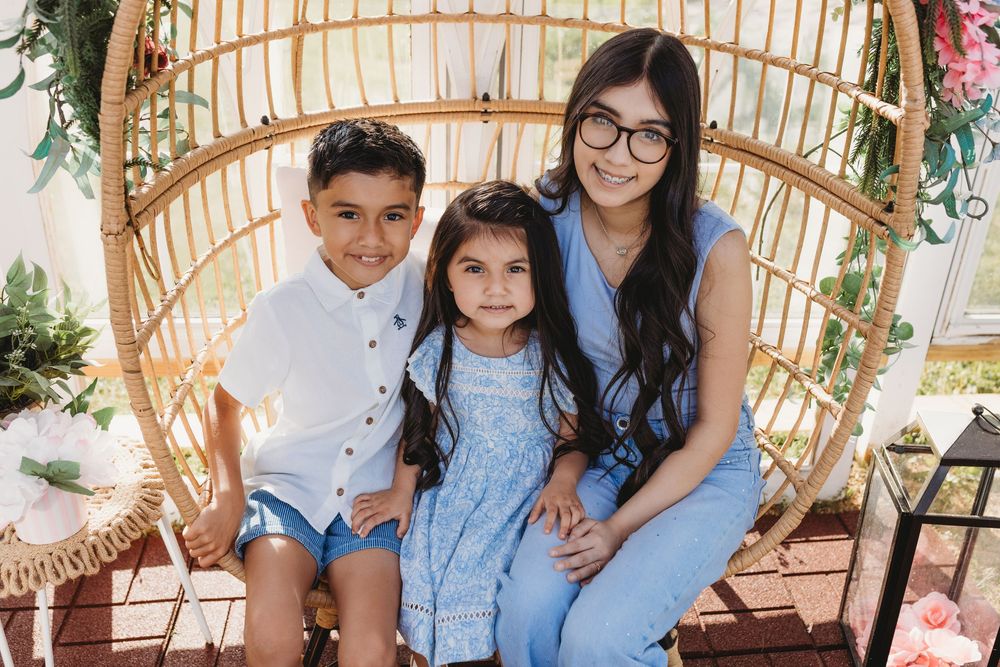 Woman with glasses posing with two young children in a rattan egg chair with floral decor.