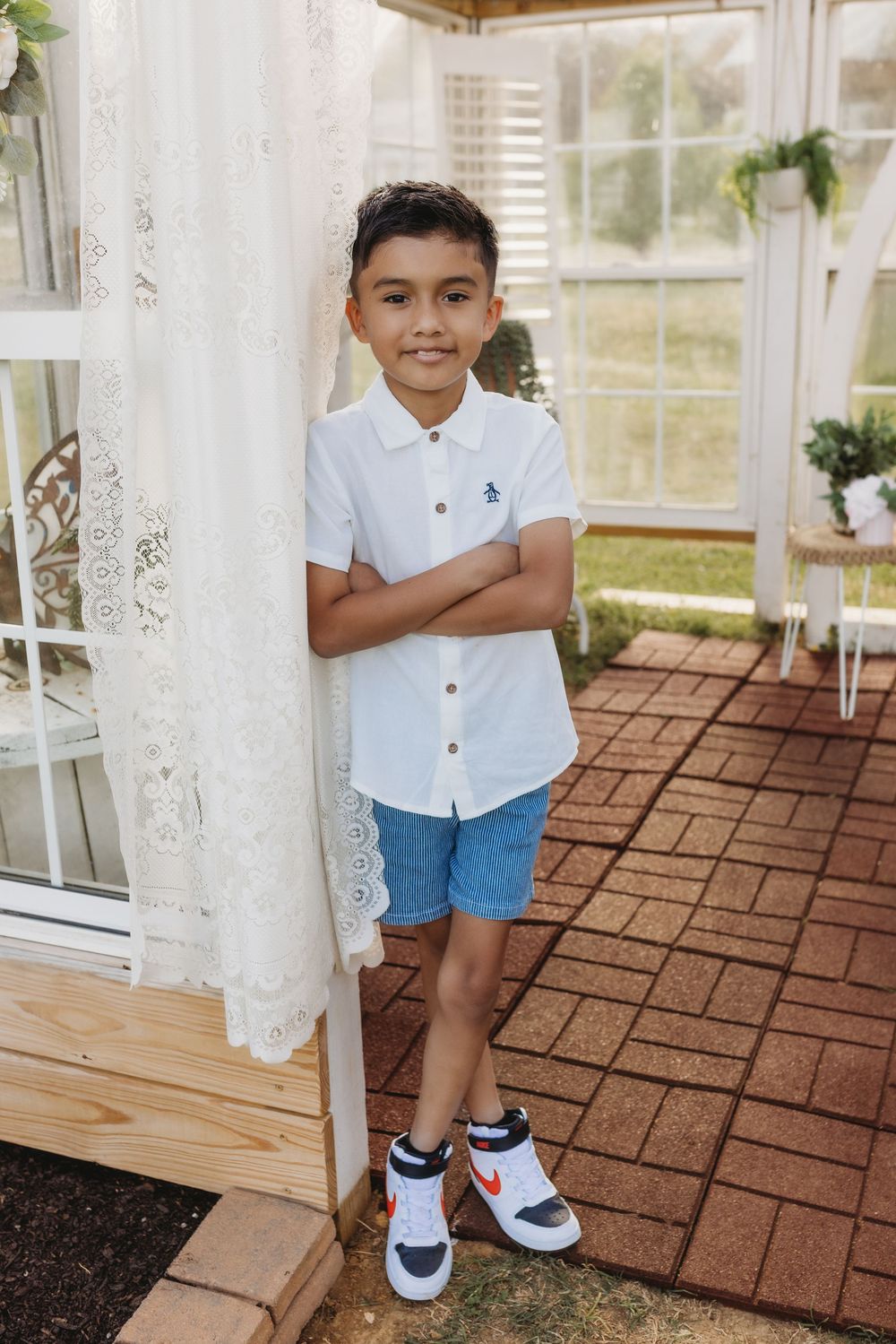 A young boy in a white polo shirt and denim shorts stands confidently with arms crossed on a brick porch.