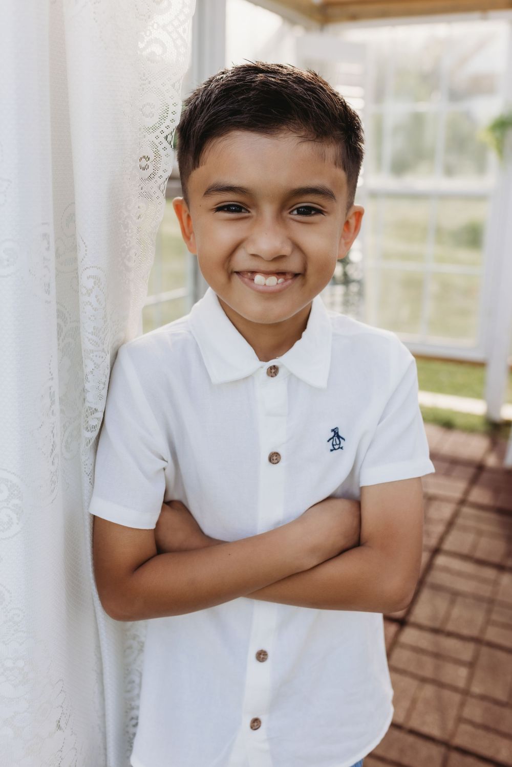 Happy boy in white Penguin polo shirt standing with arms crossed against white curtain.