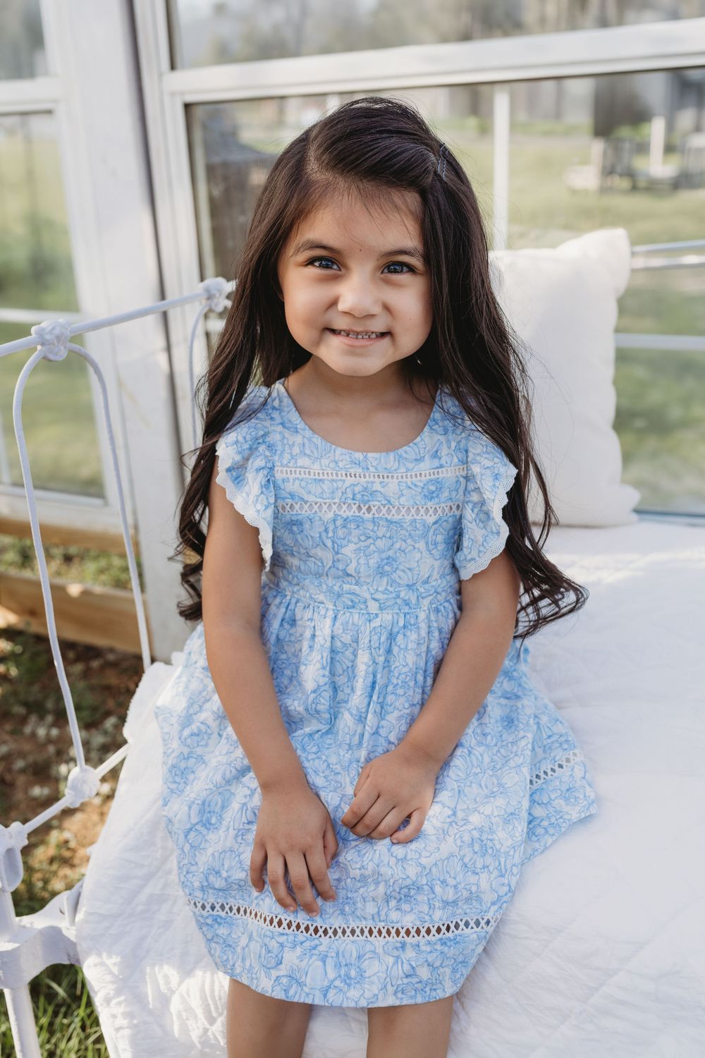 A little girl with long dark hair smiles sweetly in a blue floral dress while sitting on a white bed.