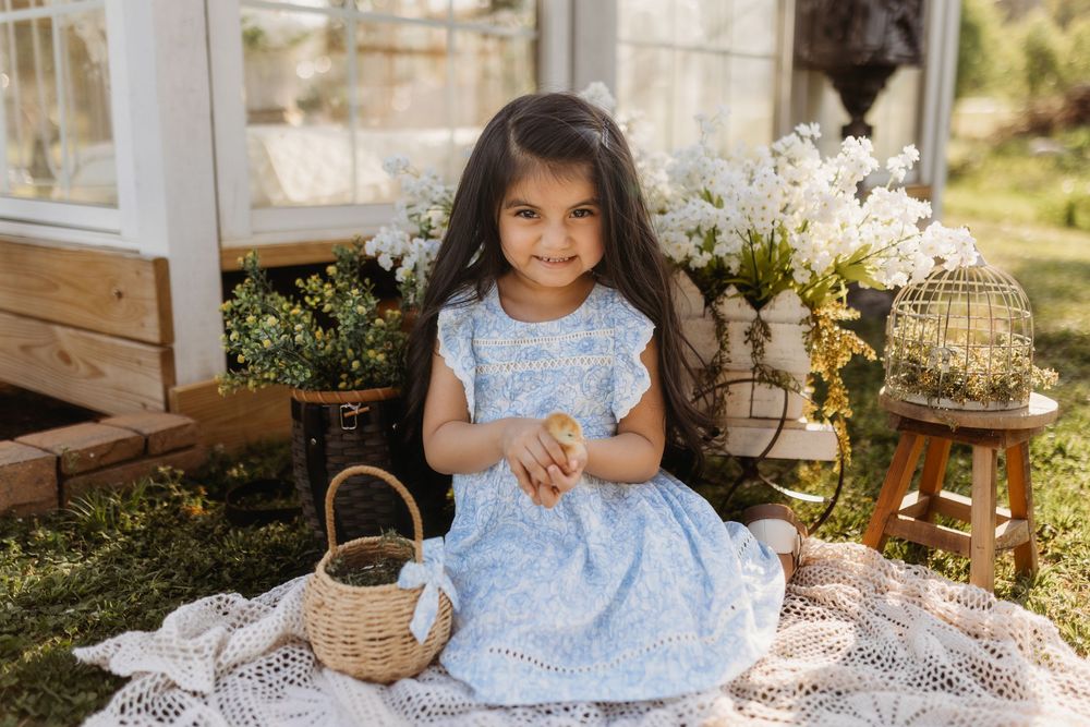 Young girl in blue floral dress sits on lace blanket with wicker basket, smiling outdoors near white flowers.