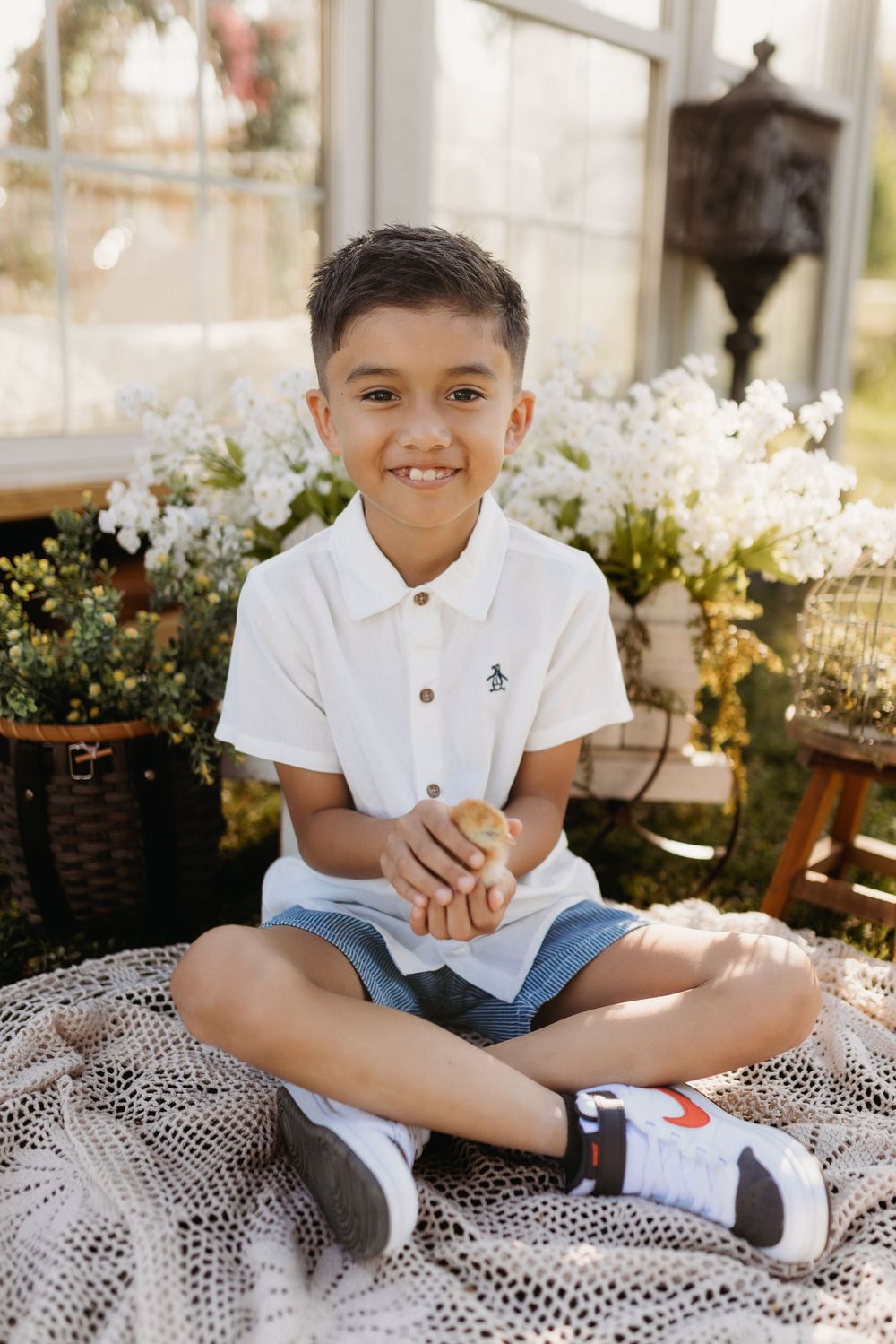 Smiling boy in white polo shirt and denim shorts sitting cross-legged on lace blanket with white flowers behind him.