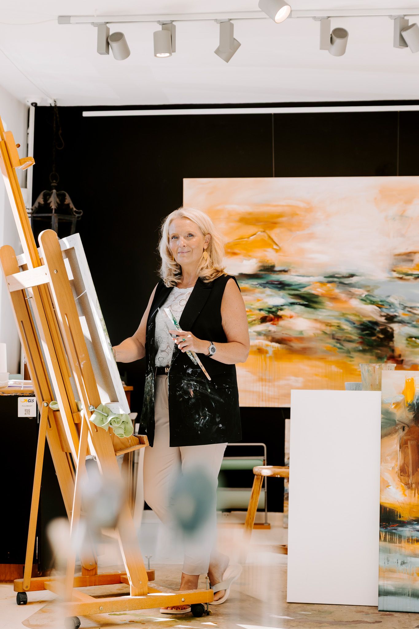 Kunstner står i sit atelier ved siden af et staffeli og holder pensler, med store abstrakte orange malerier i baggrunden. FotografMiddelfart