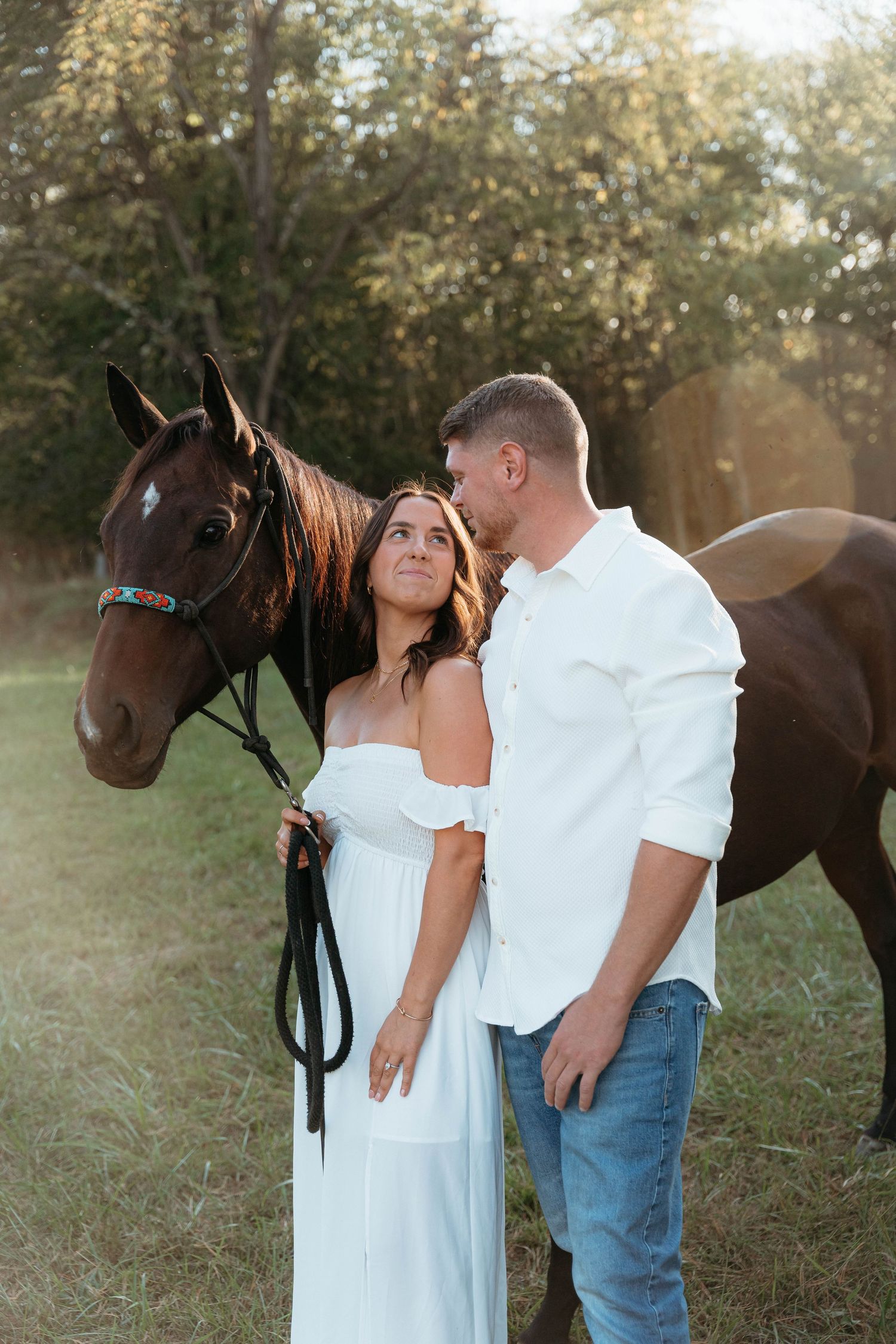 Romantic couple poses with a brown horse in a sunlit field during a photo session.