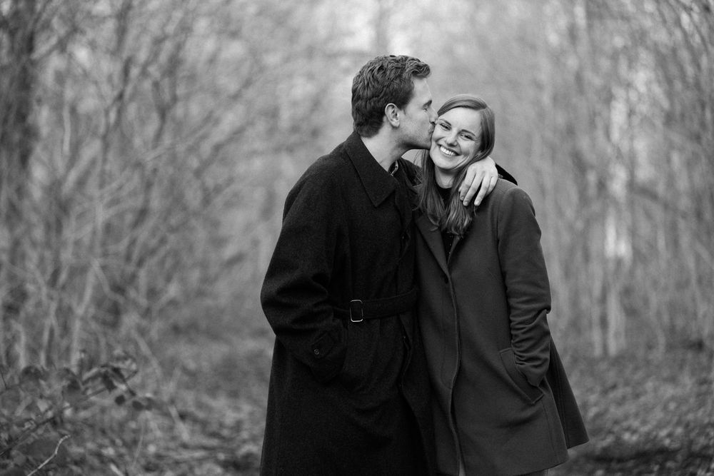 A romantic black and white photo of a couple in winter coats sharing an intimate moment on a foggy wooded path.