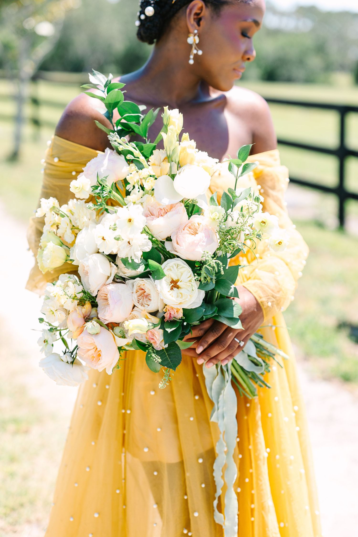 A person in a flowing yellow tulle dress holds a delicate bouquet of white and blush flowers against a pastoral backdrop.