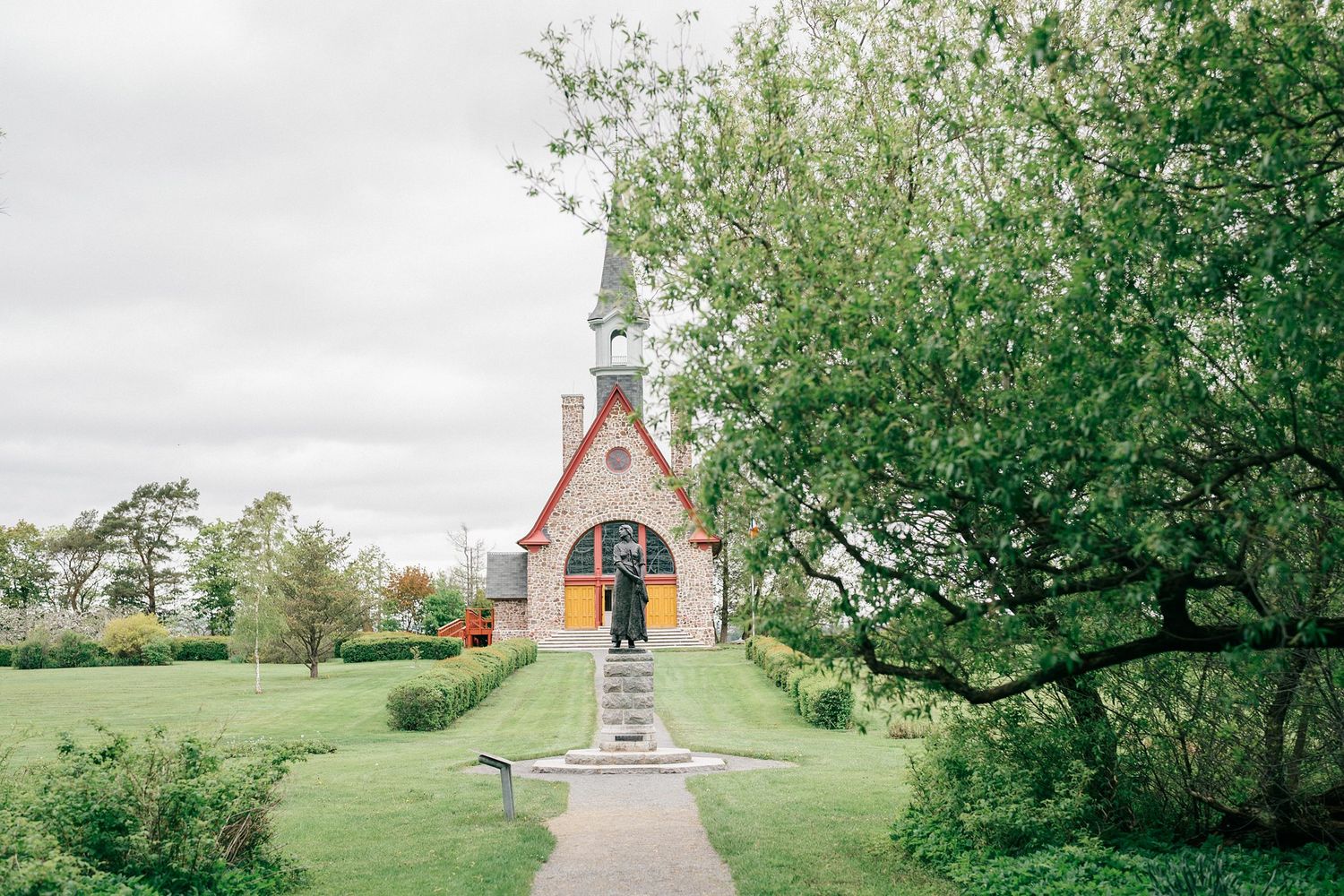 A rustic brick chapel sits at the end of a tree-lined path in a serene countryside setting.
