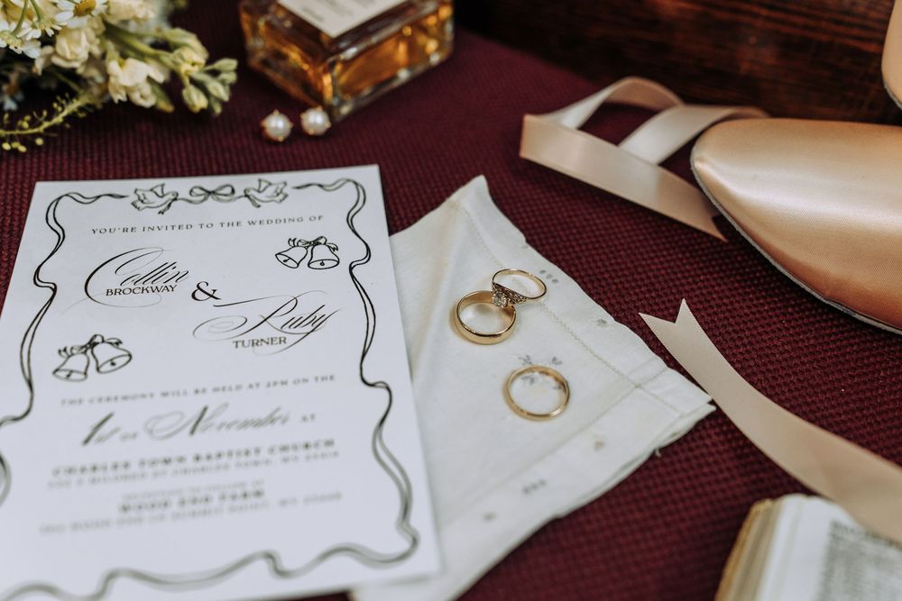 Wedding invitation, gold rings, and bridal shoes displayed on burgundy surface.