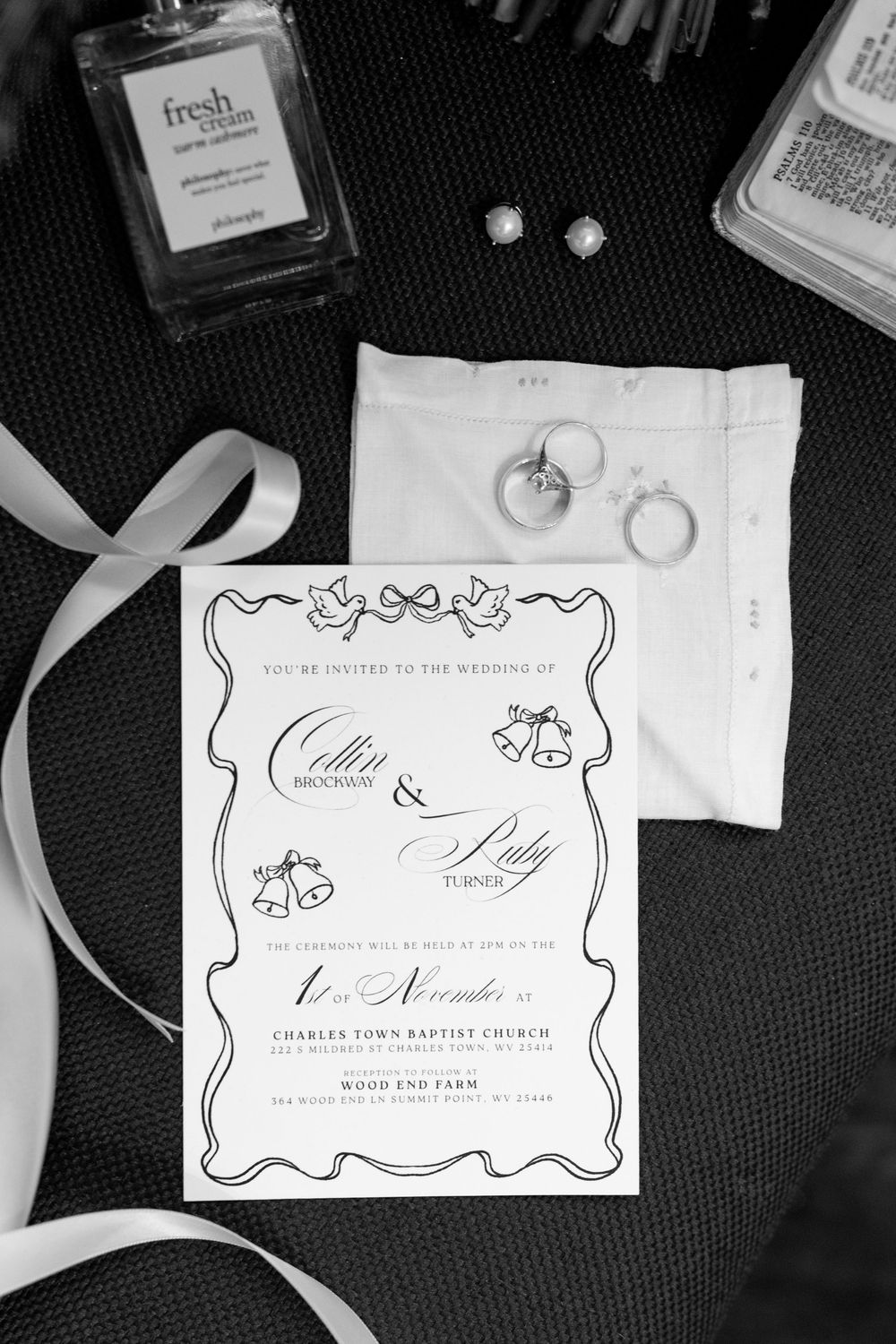 Black and white flat lay of wedding invitation, rings, earrings, and perfume bottle.