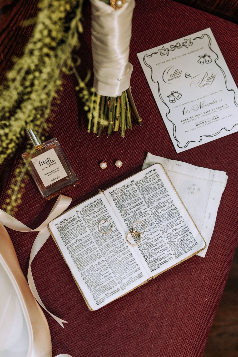 Wedding flat lay featuring open Bible with gold rings, Fresh perfume, pearl earrings, and elegant invitation on burgundy surface.