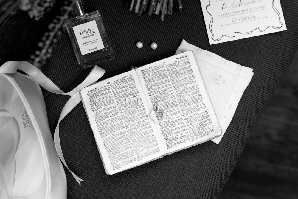 Black and white flat lay of open Bible with wedding rings, perfume bottle, and invitation on dark surface.
