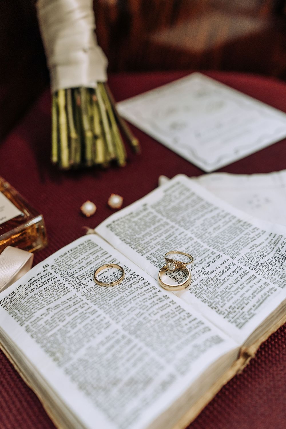 Close-up of two gold wedding bands resting on open Bible pages with bouquet and stationery in background.