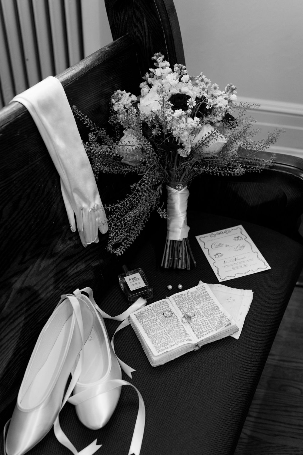 Black and white bridal flat lay with floral bouquet, white gloves, ballet shoes, Bible, and rings on church pew.