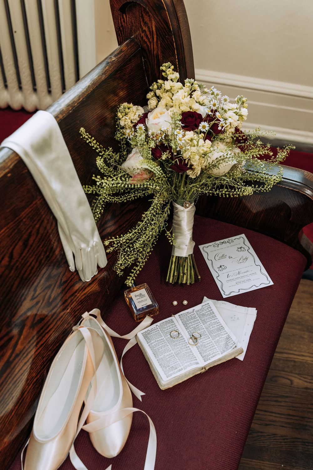Bridal details on church pew including floral bouquet, white gloves, ballet shoes, Bible with rings, and perfume.