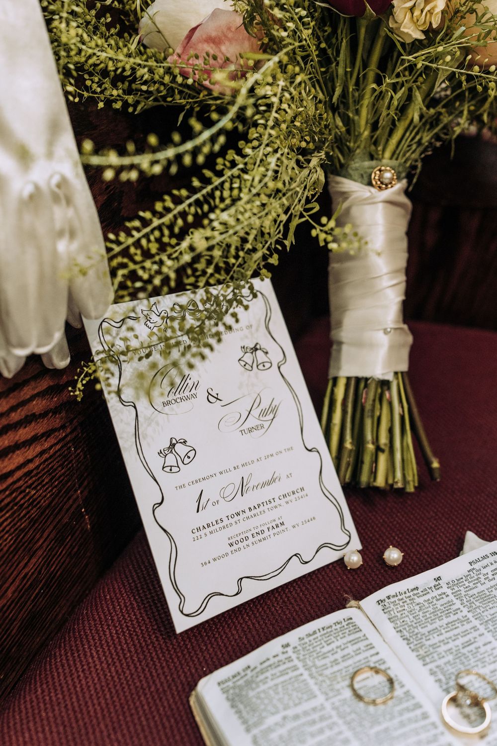Elegant wedding invitation for Collin and Ruby displayed beside floral bouquet and open Bible with gold rings.