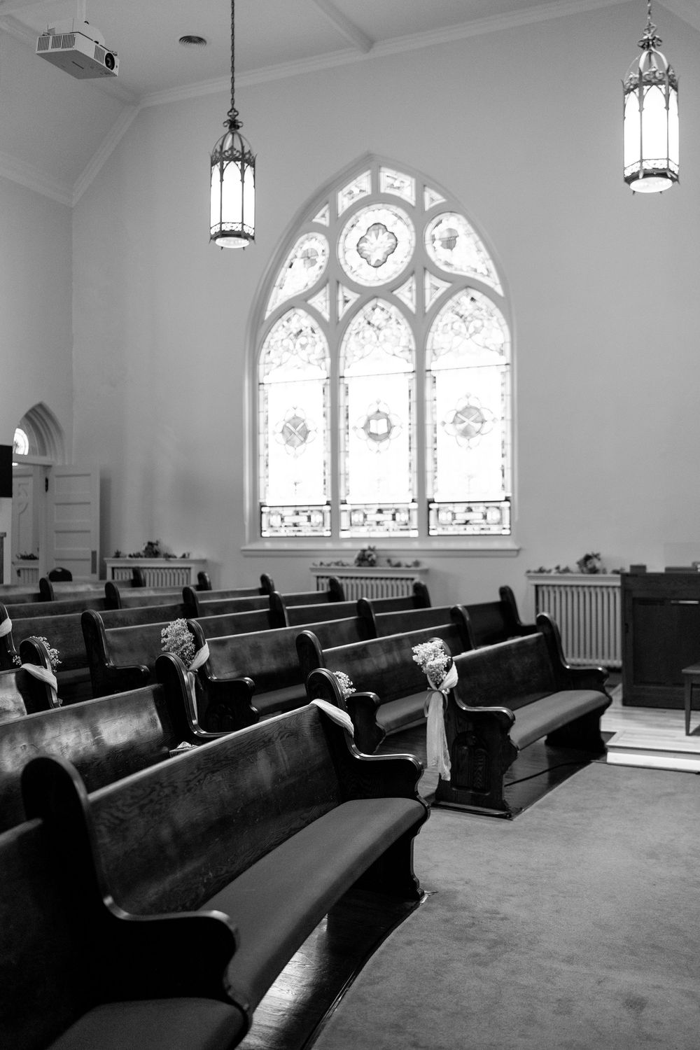 Black and white interior of traditional church with wooden pews, stained glass window, and hanging lantern chandeliers.