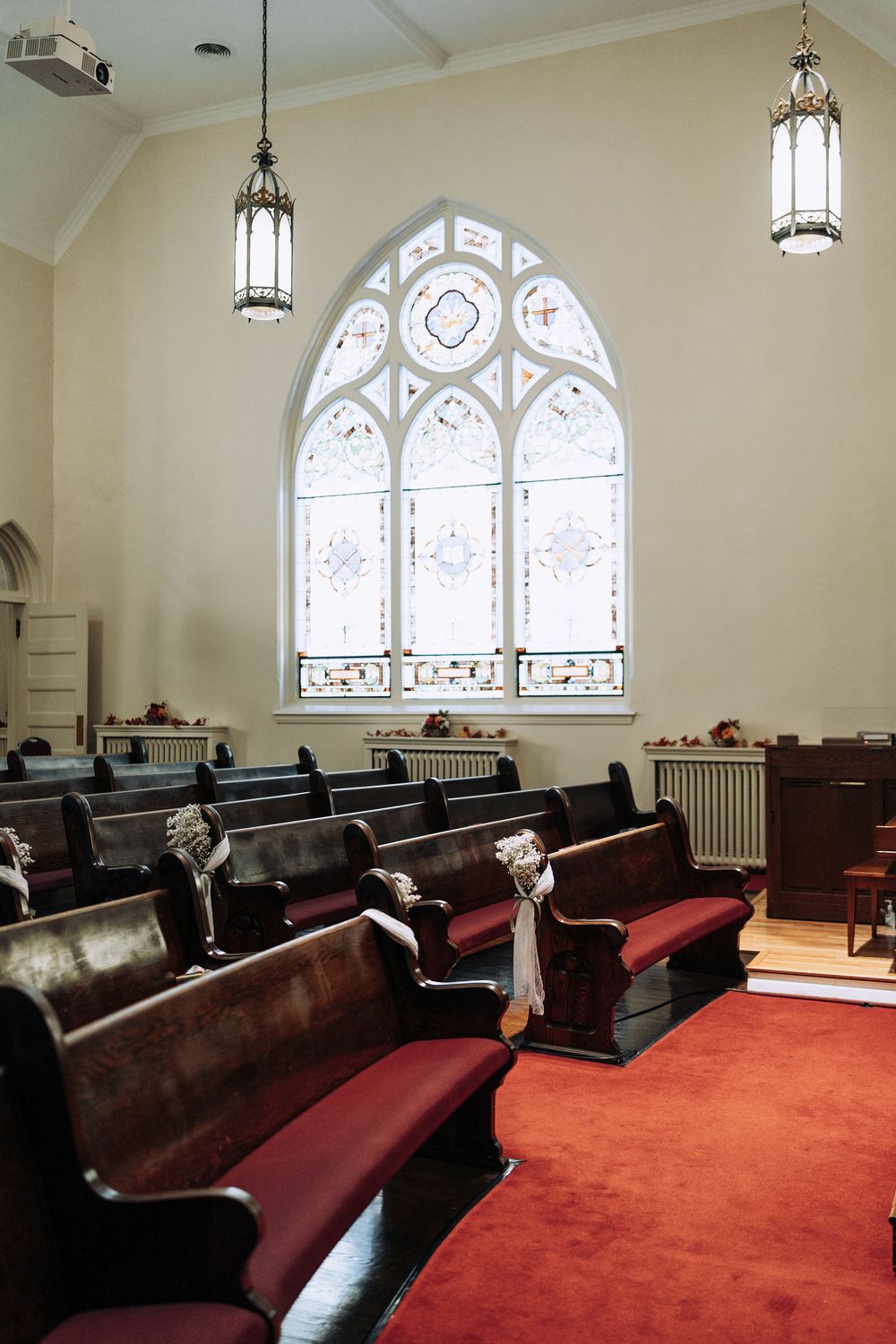 Classic church interior with red carpet aisle, dark wooden pews with burgundy cushions, and Gothic stained glass window.
