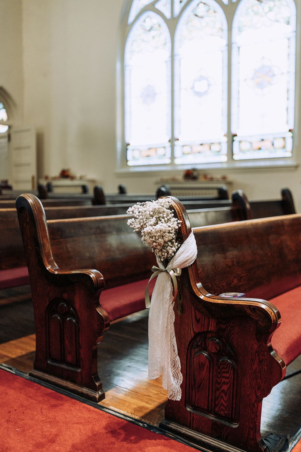 Baby's breath bouquet tied with white ribbon decorating end of wooden church pew with stained glass windows behind.