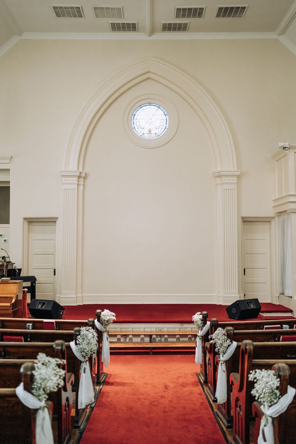 Church ceremony aisle with red carpet, wooden pews decorated with white floral arrangements, leading to arched altar wall.