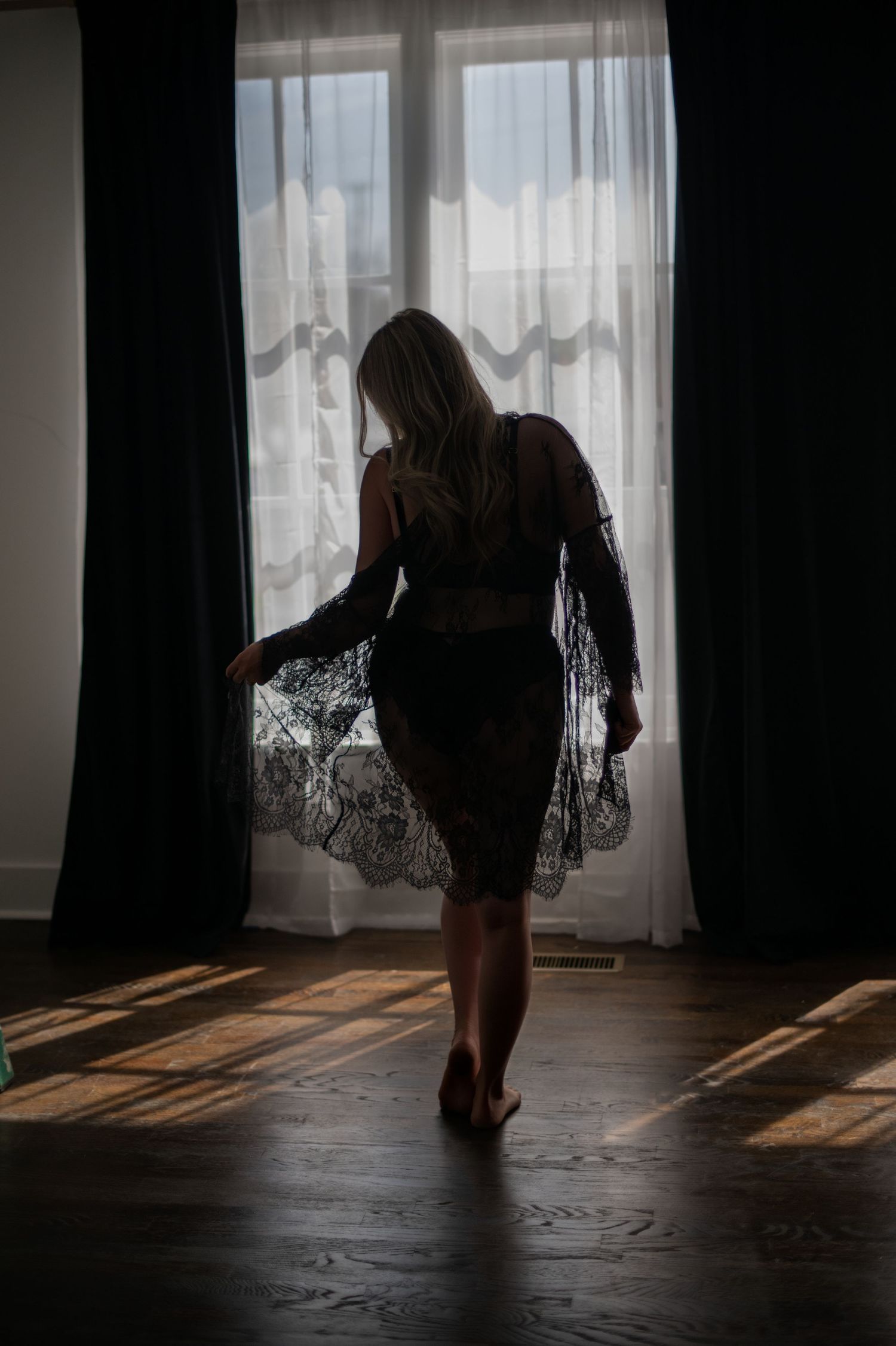 A silhouetted figure stands in a dimly lit room with sheer curtains casting dramatic shadows across the wooden floor.