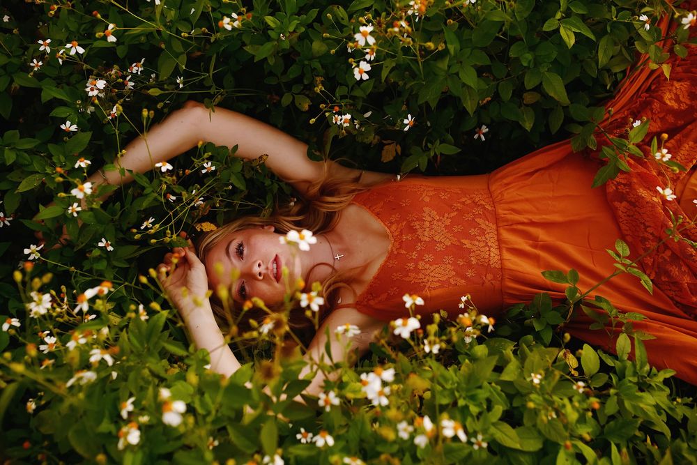 Someone in a vibrant orange dress lies dreamily among flowering bushes in a garden setting.