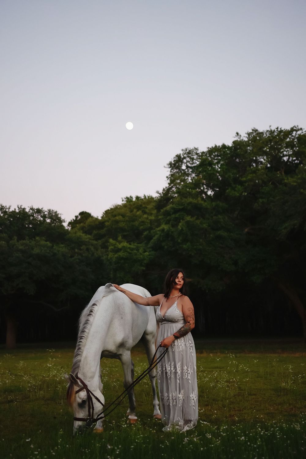 A white horse grazes in a moonlit meadow beside a person wearing a flowing white dress at dusk.
