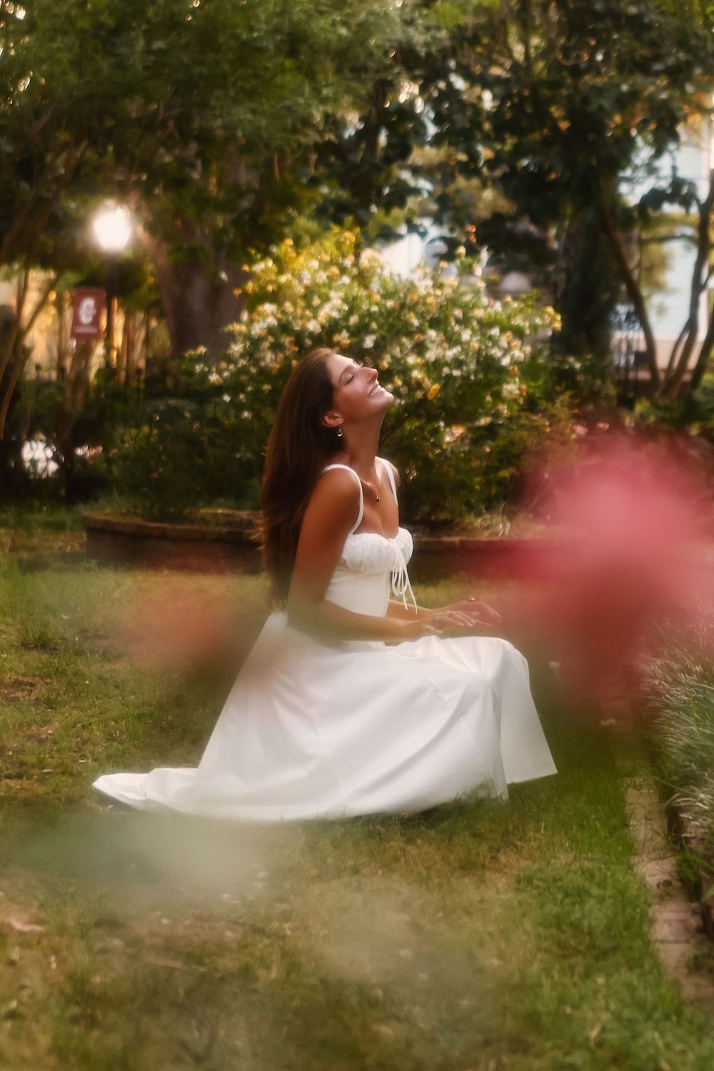 A person in a flowing white dress reclines gracefully on grass in a dreamy garden setting during golden hour.