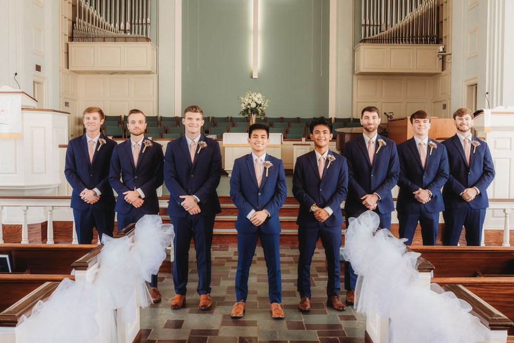 A wedding party in navy suits stands in formation at the altar of a church with white pew decorations.