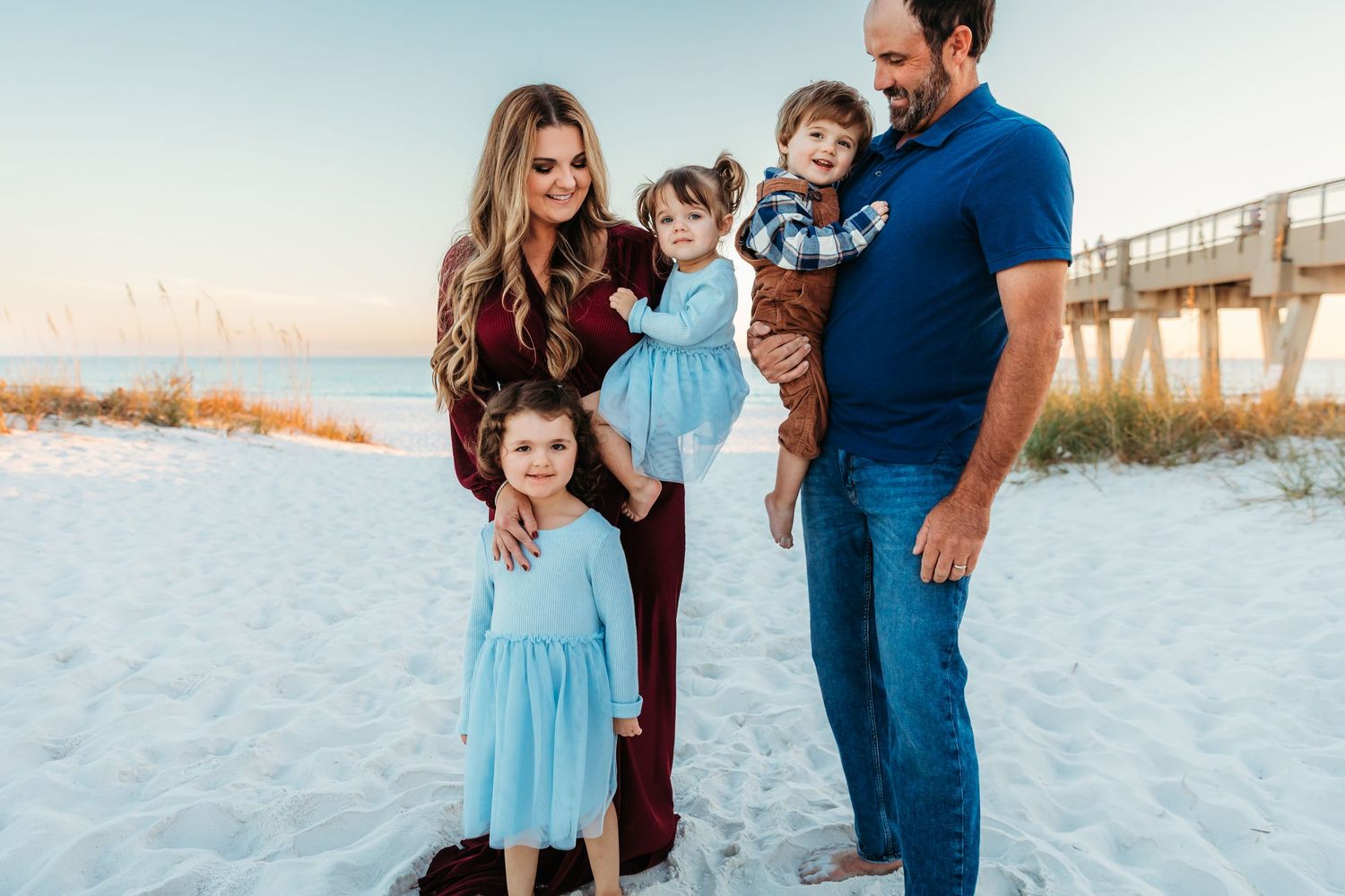 Family wearing coordinating blue and burgundy outfits poses together on white sandy beach at sunset.