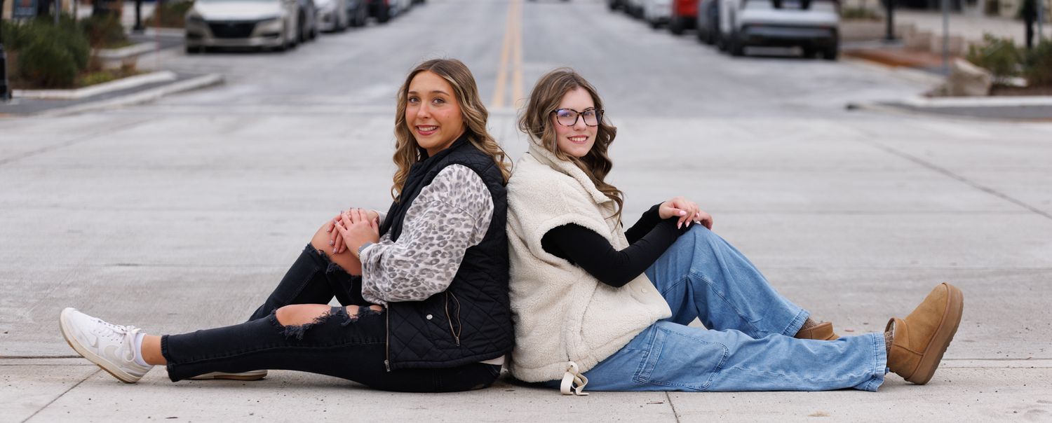 Two high school senior girls posed sitting back to back in downtown scene.