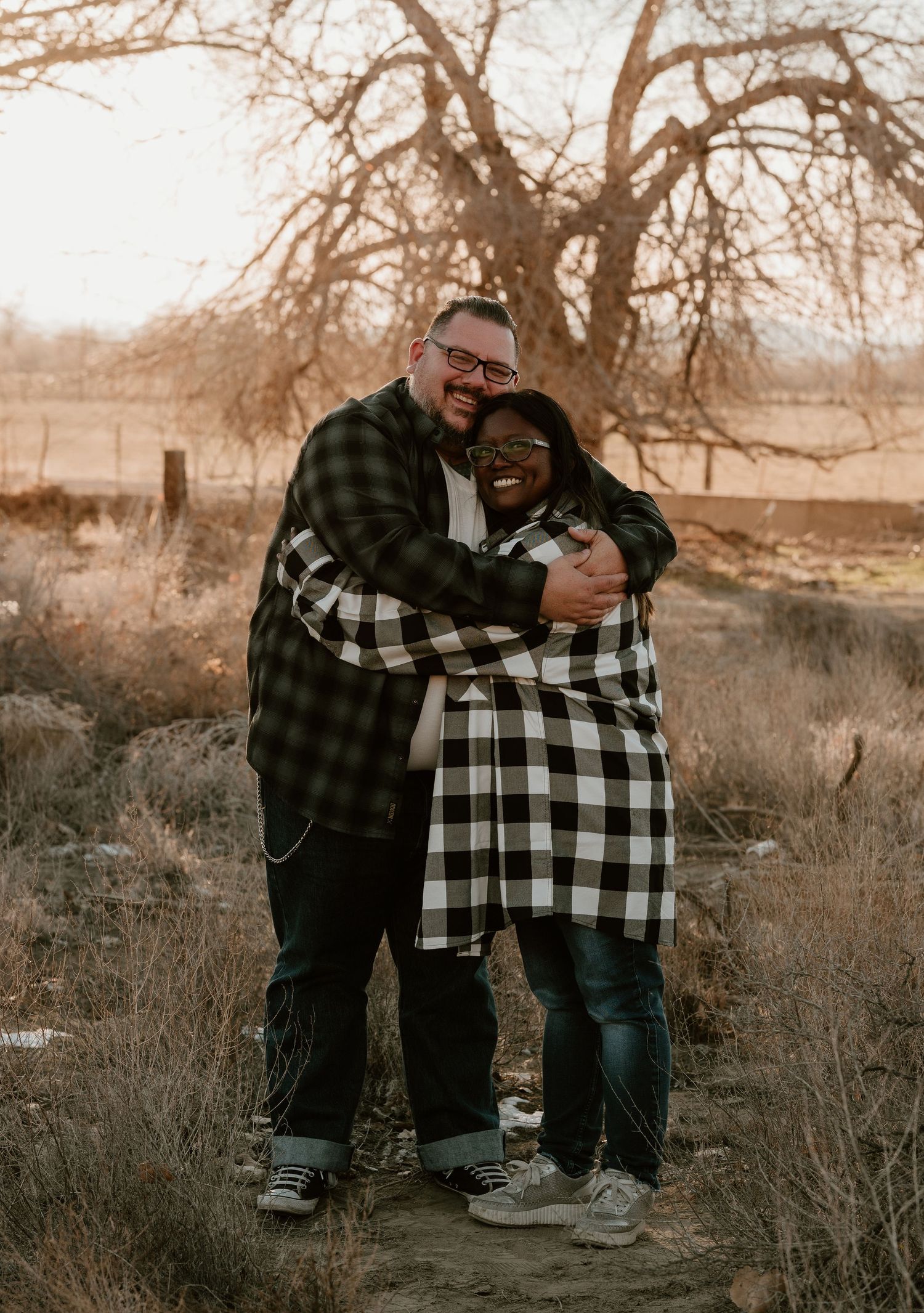 Two people in plaid shirts share a tender embrace outdoors during golden hour in a rustic autumn setting.