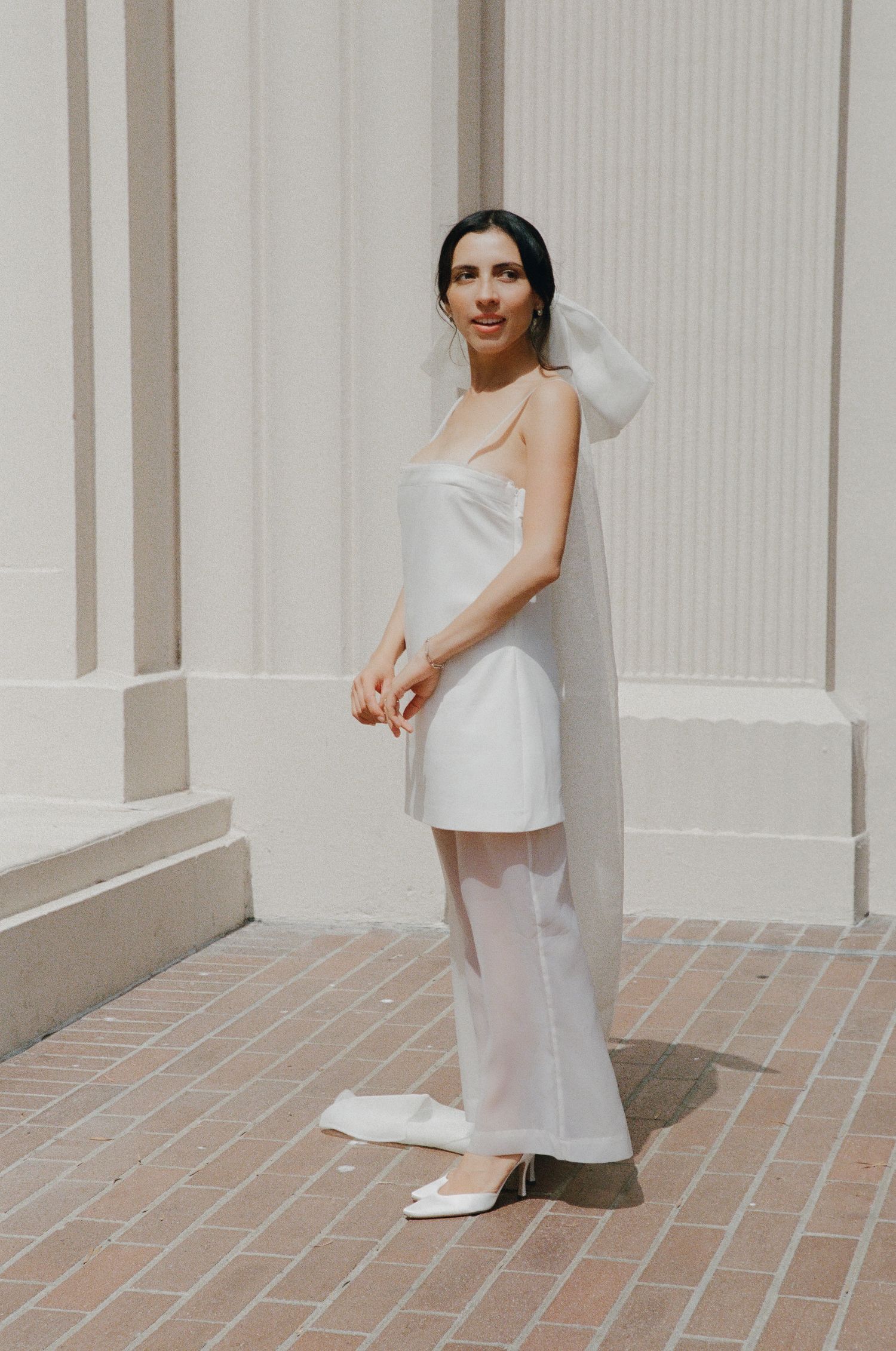Woman in white dress poses elegantly against architectural columns and brick floor.
