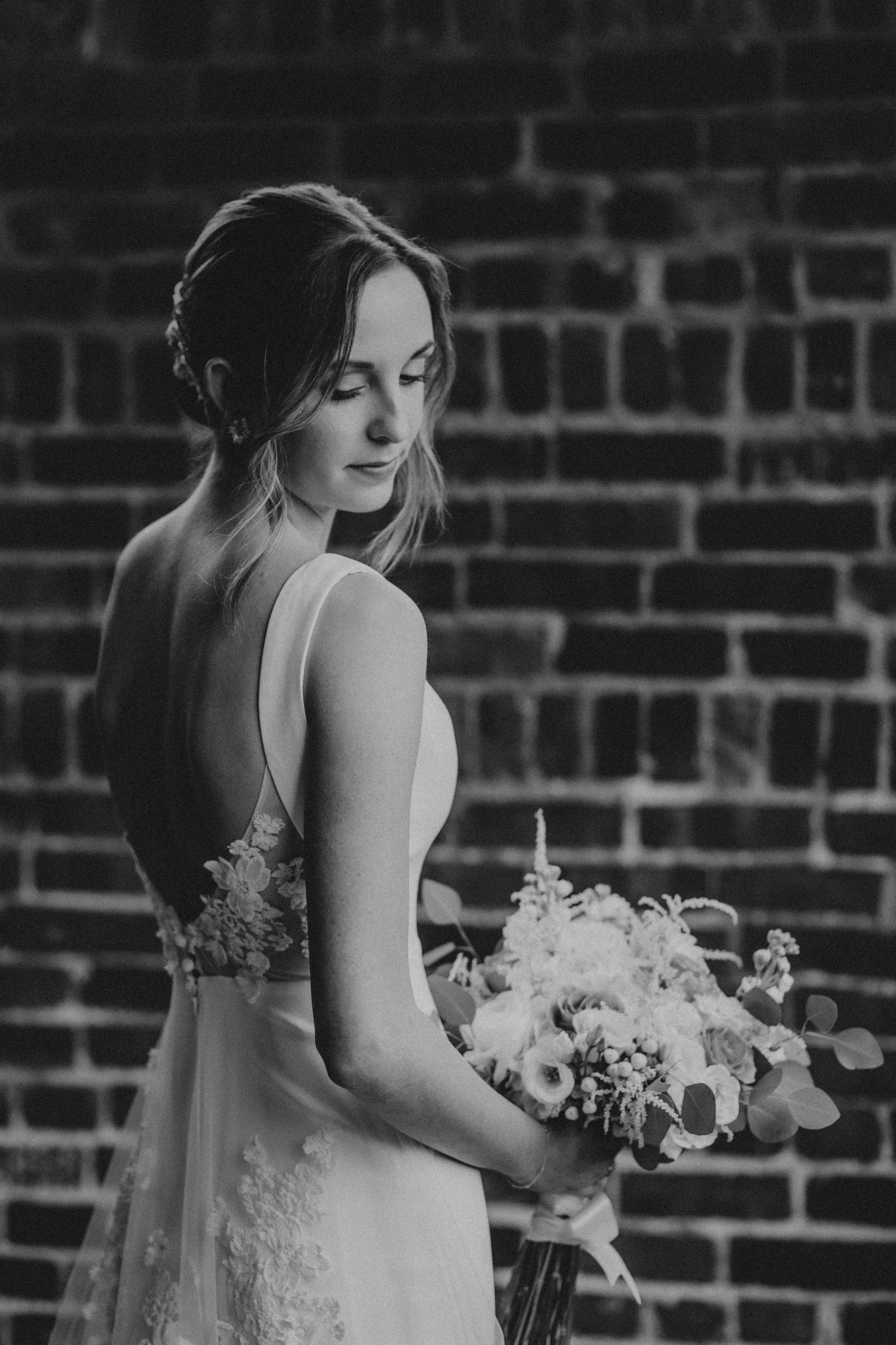 Black and white photo of a bride in a lace wedding dress against a brick wall with flowers.