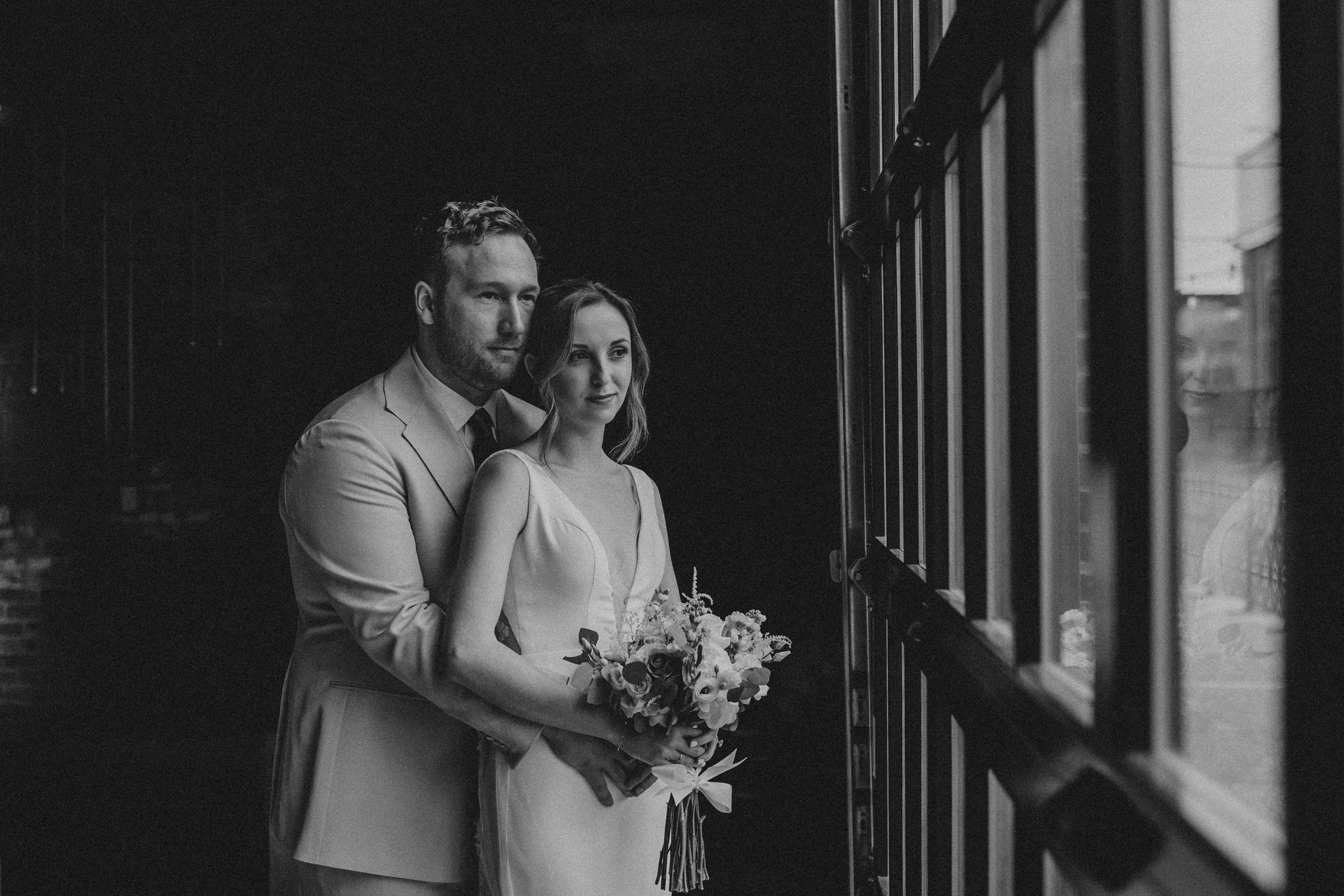 A black and white wedding portrait of a couple standing by a window, with the bride holding a floral bouquet.