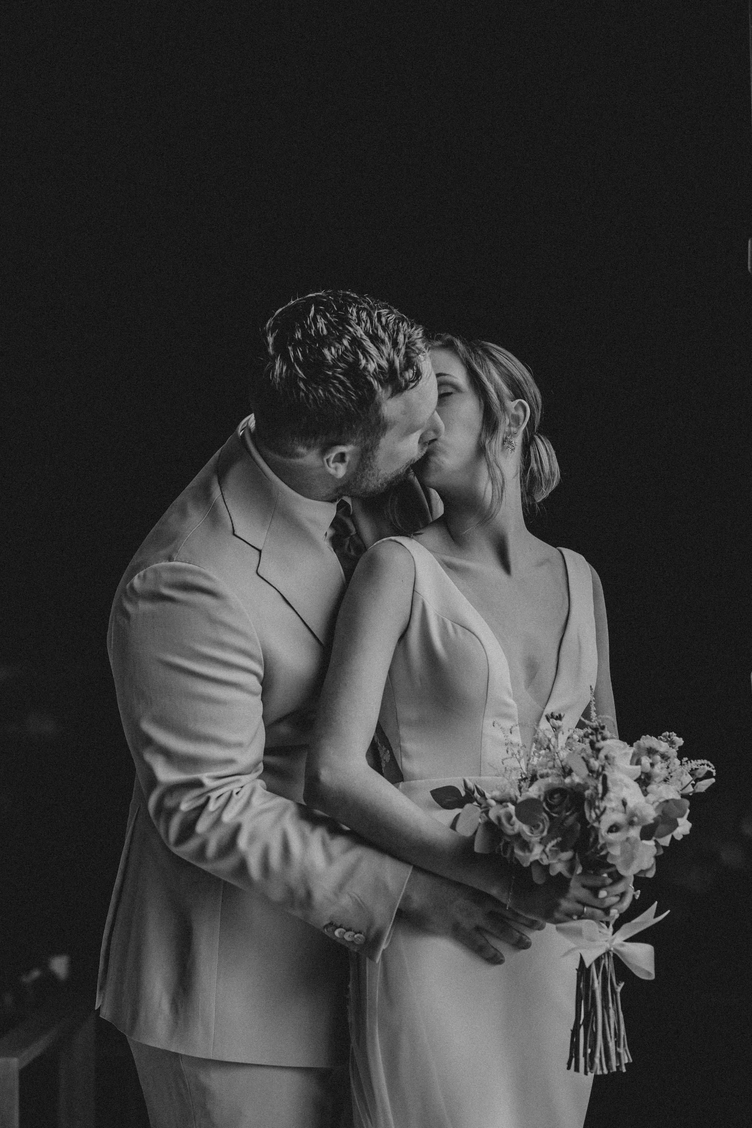 A romantic black and white wedding photo of a couple sharing an intimate kiss while holding a floral bouquet.