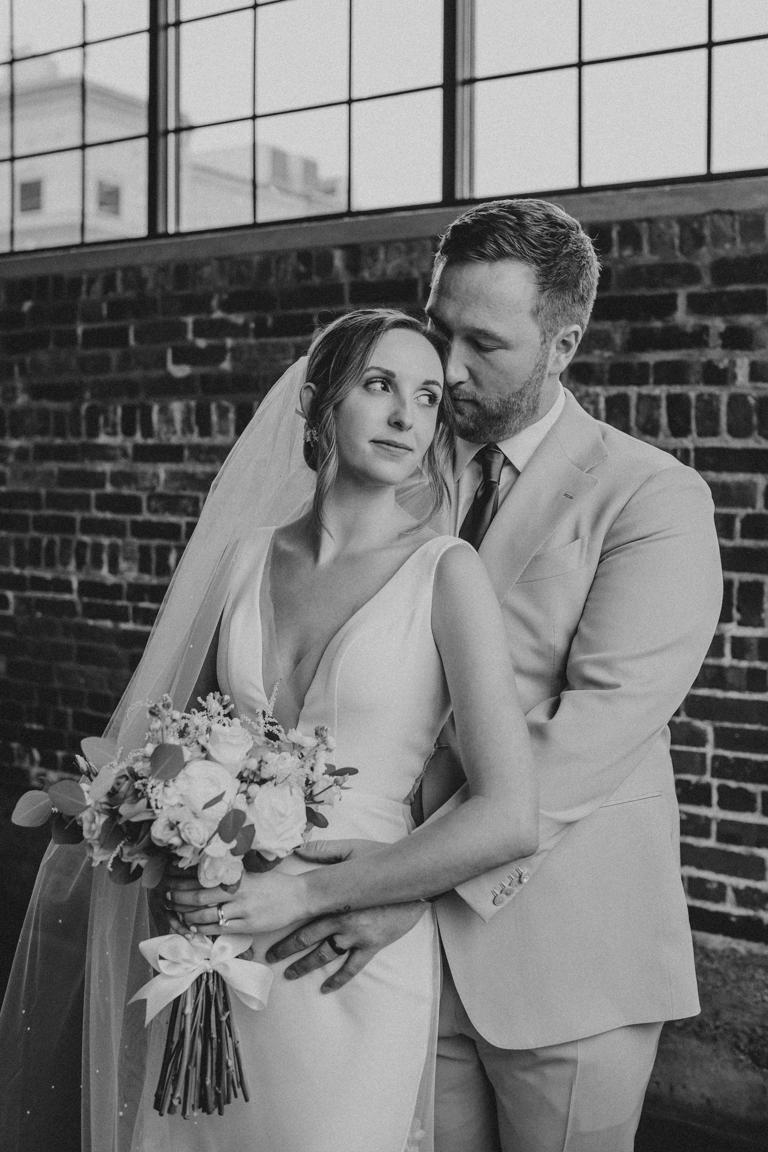 Black and white wedding photo of a couple embracing against a brick wall, bride holding a floral bouquet.