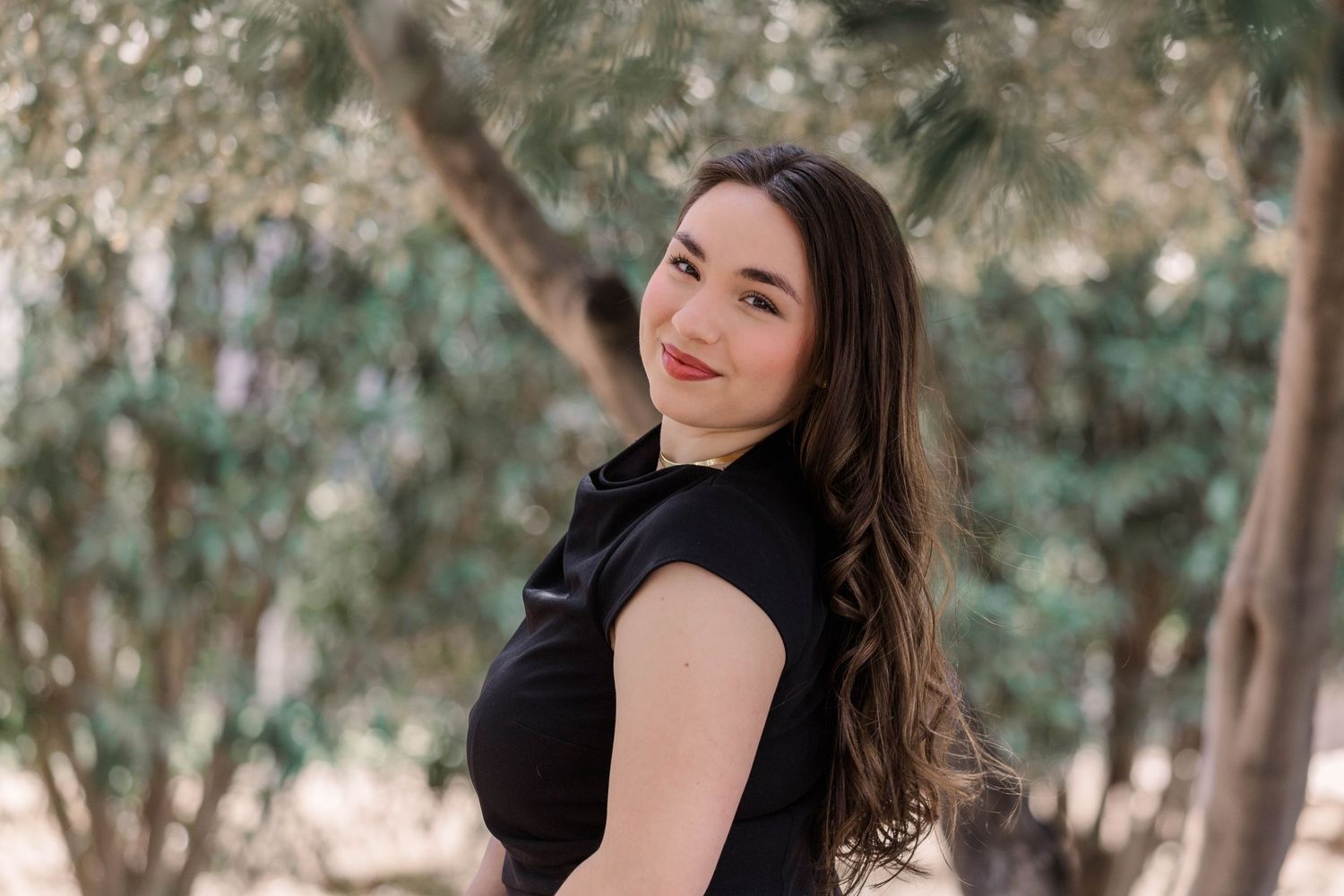 A person in a black dress poses gracefully near a tree trunk in a natural outdoor setting.