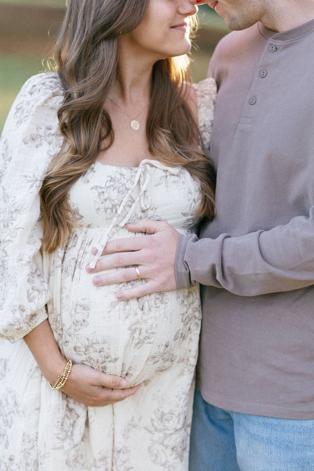 A couple embraces in a romantic maternity photoshoot, wearing a floral dress and grey sweater against a sunlit backdrop.