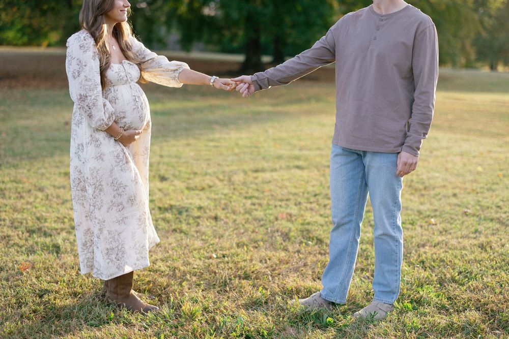 A couple holds hands in a sunlit field, with the woman wearing a white lace maternity dress.