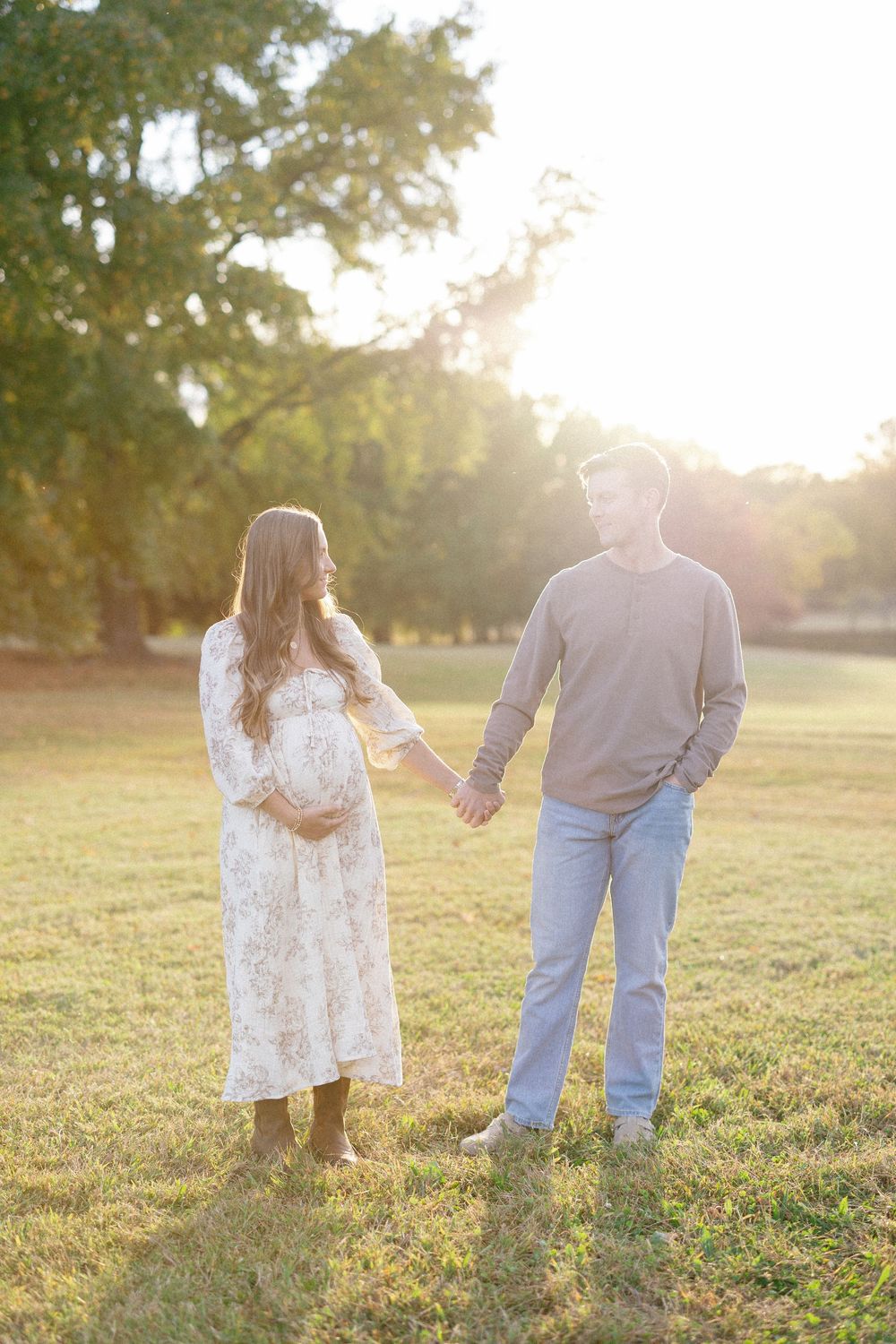 A couple holds hands while standing in a sunlit grassy field during golden hour.